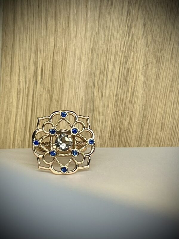 Bague Princesse en vermeil quartz fume saphirs bleus collection les pierres vue de face | 26 Passage, Créatrice de bijoux à Paris Gold floral filigree ring with blue stones around a central clear gem on a pale surface