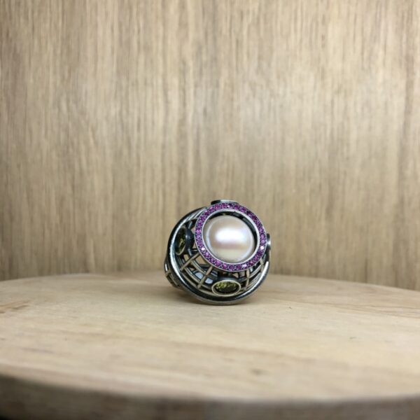 bague marie antoinette argent noir peridots ligne saphirs rose et perle eau douce creme vue de face | 26 Passage, Créatrice de bijoux à Paris Silver ring with a large central pearl surrounded by a pink gemstone halo, resting on a wooden surface.