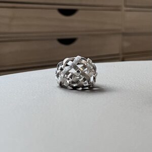 bague tressee argent vue de face Close-up of a silver braided ring resting on a pale desk, with a brown filing cabinet blurred in the background.