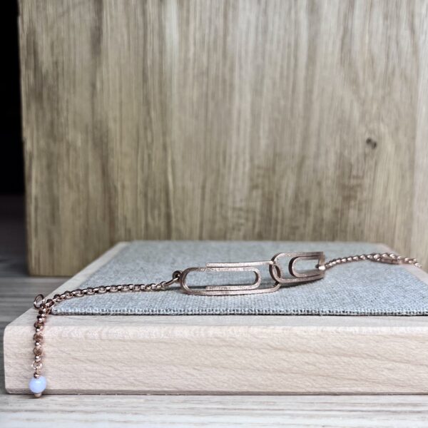 bracelet double trombone vermeil a plat | 26 Passage, Créatrice de bijoux à Paris Rose-gold chain necklace with a blue bead pendant on a light blue mat atop a wooden surface in this close-up view.