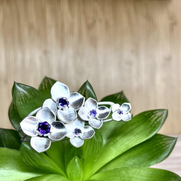 bague orthensia argent et amethystes posee sur une plante verte | 26 Passage, Créatrice de bijoux à Paris Close-up of a bouquet of metallic silver flowers with purple gemstone centers amid glossy green leaves on a wooden background.