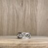 bague filaire ronde saphirs bleux collection femme vue face | 26 Passage, Créatrice de bijoux à Paris Silver ring with an intricate interlaced knot design and a small central blue gemstone on a light wooden surface.
