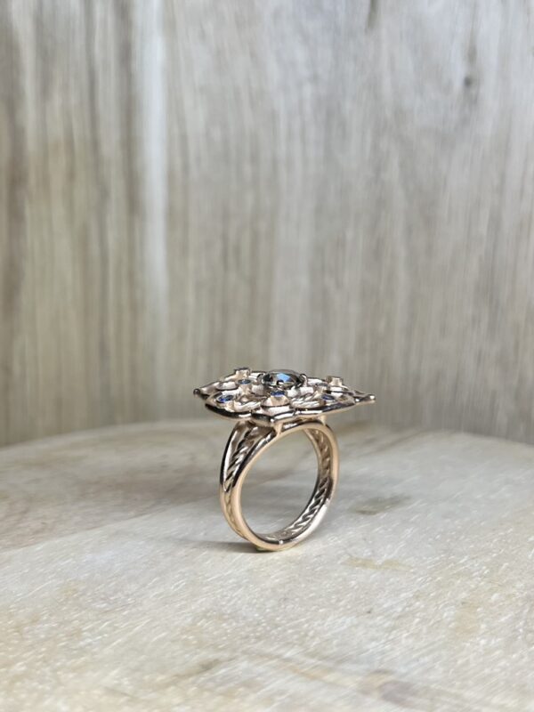Bague Princesse en vermeil quartz fume saphirs bleus collection les pierres vue profil | 26 Passage, Créatrice de bijoux à Paris Rose-gold ring with a floral cluster of blue gemstones set on a carved band, photographed on a light surface at angle view