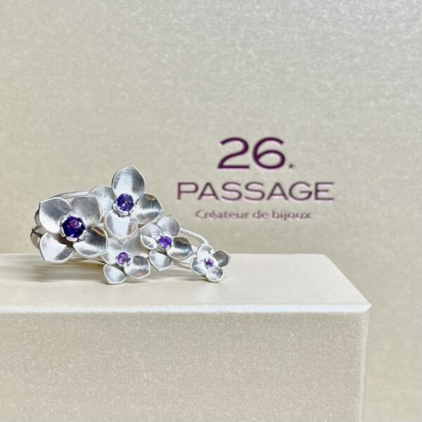 bague orthensia argent amethystes sur deux doigts posee sur ecrin | 26 Passage, Créatrice de bijoux à Paris Silver flower-adorned ring with purple gemstones resting on a white display block, logo '26 Passage' in the background.