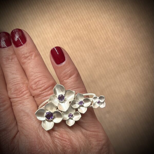 bague orthensia argent et amethystes portee deux doigts | 26 Passage, Créatrice de bijoux à Paris Hand with red nail polish wearing a silver flower bracelet with purple gemstones on the wrist/knuckles area.
