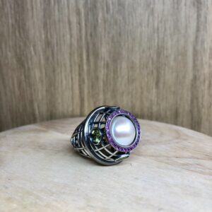 Silver lattice ring with a large central pearl surrounded by a pink gemstone halo and a small green accent stone on the side.