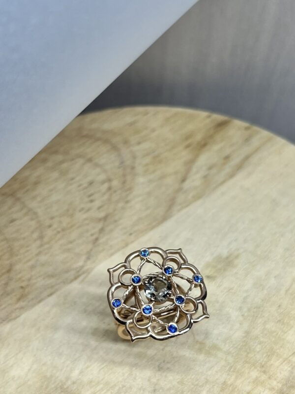 Bague Princesse en vermeil quartz fume saphirs bleus collection les pierres vue dessus | 26 Passage, Créatrice de bijoux à Paris Intricate flower-shaped ring with blue stones at the petal edges and a central gray gemstone on a light surface.