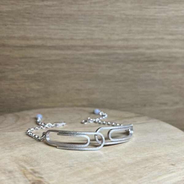 bracelet double trombone argent a plat | 26 Passage, Créatrice de bijoux à Paris Silver bracelet with interlocking rectangular links resting on a light wooden surface, sparkly finish visible at edges.