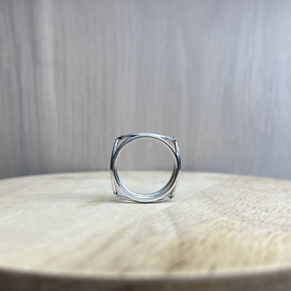 bague filaire carree arrondi saphir parme collection femme vue cote | 26 Passage, Créatrice de bijoux à Paris Close-up of a hollow silver ring setting standing on a light wooden tabletop with a blurred gray background.