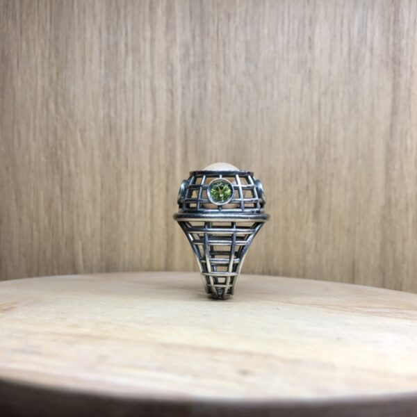 bague marie antoinette argent noir peridots ligne saphirs rose et perle eau douce creme vue de cote | 26 Passage, Créatrice de bijoux à Paris Metal lattice-style ring with a round green gemstone, displayed on a light wooden surface.