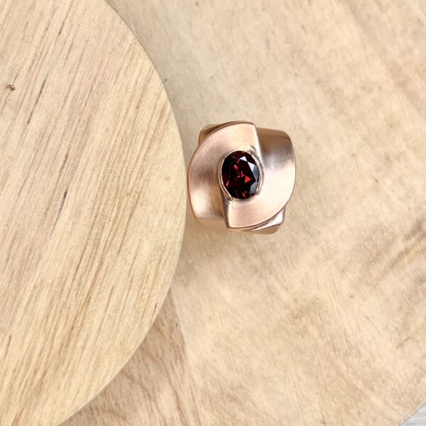 Bague twist vermeil grenat collection femme | 26 Passage, Créatrice de bijoux à Paris Rose-gold colored ring with a chunky, squared band and a faceted deep red gemstone set in a low bezel, resting on a light wooden surface.