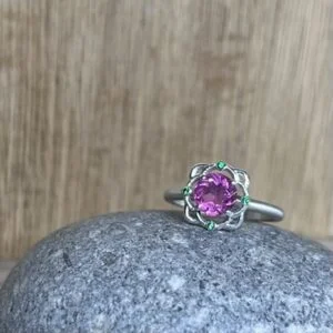 Bague mini princesse argent tourmaline rose grenats tsavorite Silver ring with a pink gemstone centerpiece, accented by small green stones, resting on a gray rock with a wooden background.