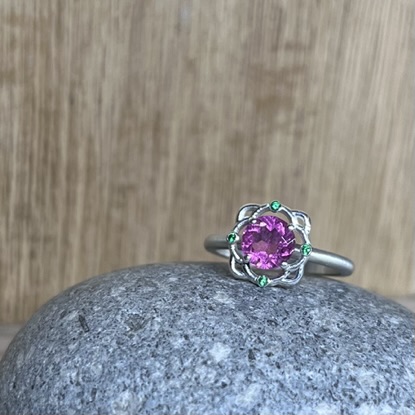 Silver ring with a pink gemstone centerpiece, accented by small green stones, resting on a gray rock with a wooden background.