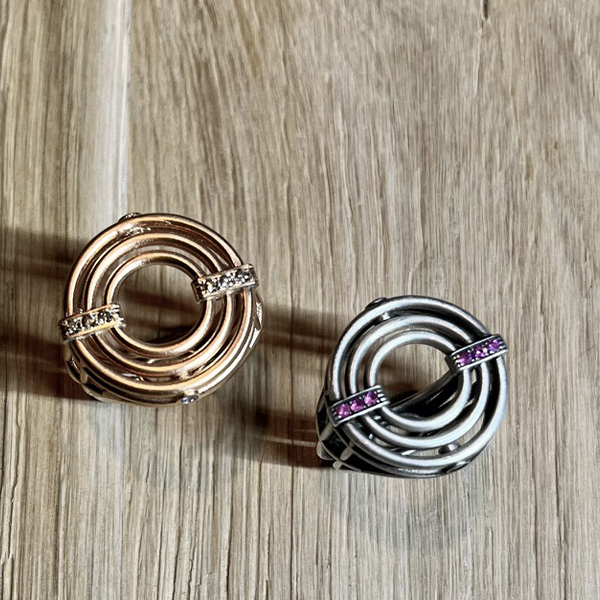 bague meringue vermeil et argent vue dessus | 26 Passage, Créatrice de bijoux à Paris Two circular rings with intertwined inner loops, one rose gold with clear stones and one silver with purple stones, on a wooden surface.