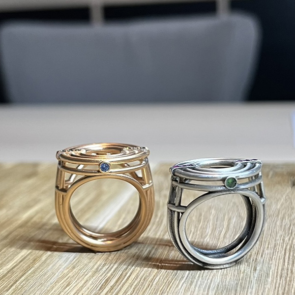 bague meringue vermeil vue cote | 26 Passage, Créatrice de bijoux à Paris Rose gold ring with blue gem and intricate openwork beside a silver ring with green gem on a wooden surface.