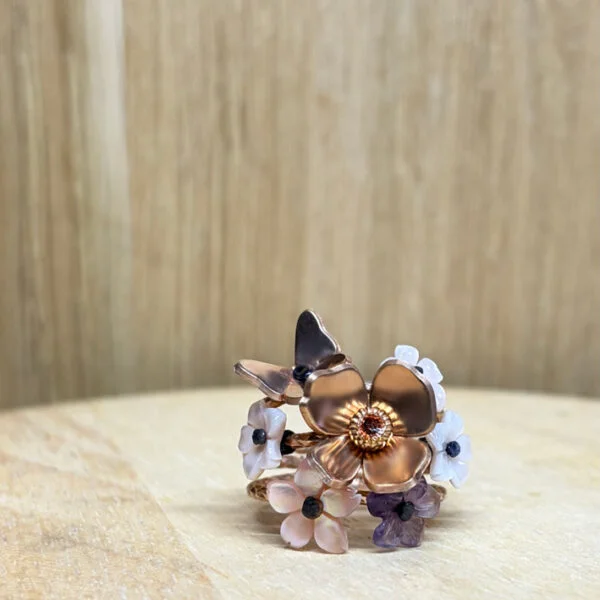 bague prairie vermeil nacre fleur amethyste | 26 Passage, Créatrice de bijoux à Paris Copper flower ring with a central gem and smaller white, pink, and purple blossoms arranged around it on a light wooden surface.