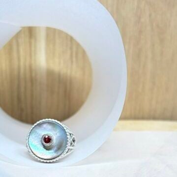 chevaliere nacre saphir sonkia vue face | 26 Passage, Créatrice de bijoux à Paris Silver ring with a red gemstone set in a decorative bezel, displayed inside a white curved form on a wooden background.