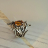 bague double branche melange vermeil argent noir citrine madere vue profil | 26 Passage, Créatrice de bijoux à Paris Ring with an oval orange gemstone held by intertwining silver branch-like prongs on a light marble surface.
