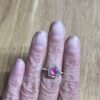 bague mini princesse argent tourmaline rose grenats tsavorite portee | 26 Passage, Créatrice de bijoux à Paris Close-up of a hand wearing a silver ring with a large pink gemstone flanked by small green stones, resting on a wooden surface.