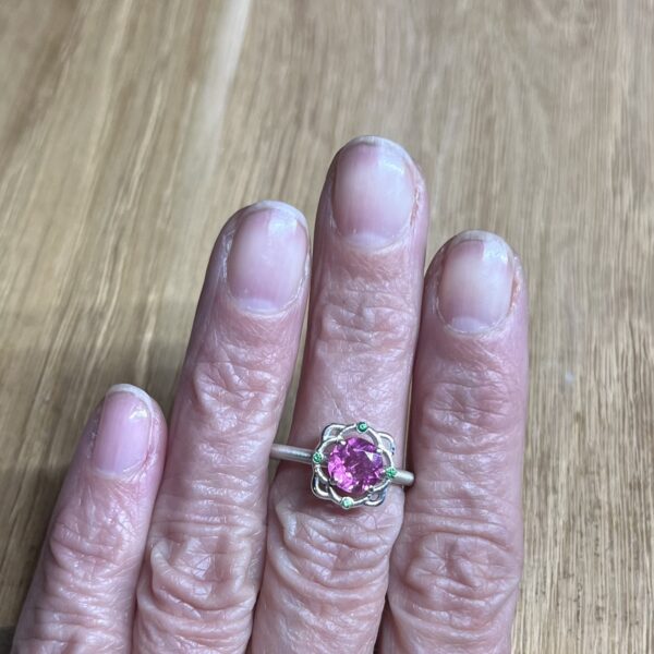 bague mini princesse argent tourmaline rose grenats tsavorite portee | 26 Passage, Créatrice de bijoux à Paris Close-up of a hand wearing a silver ring with a large pink gemstone flanked by small green stones, resting on a wooden surface.