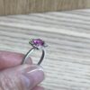 bague mini princesse argent tourmaline rose grenats tsavorite profil | 26 Passage, Créatrice de bijoux à Paris Silver ring with an oval pink-purple gemstone held between someone's fingers, displayed against a wooden background, showing a halo setting with small side stones