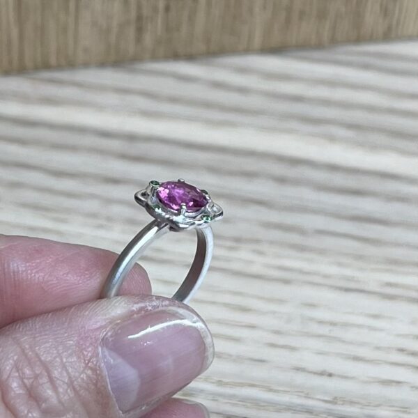 bague mini princesse argent tourmaline rose grenats tsavorite profil | 26 Passage, Créatrice de bijoux à Paris Silver ring with an oval pink-purple gemstone held between someone's fingers, displayed against a wooden background, showing a halo setting with small side stones