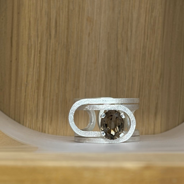 bague trombone argent quartz fume ovale vue face | 26 Passage, Créatrice de bijoux à Paris Silver ring with an oval smoky quartz gemstone set in a geometric open frame, resting on a light wood surface.