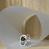 bague trombone argent quartz fume ovale vue trois quart | 26 Passage, Créatrice de bijoux à Paris Silver ring with a large dark gemstone, resting on a curved white paper backdrop on a wooden surface.