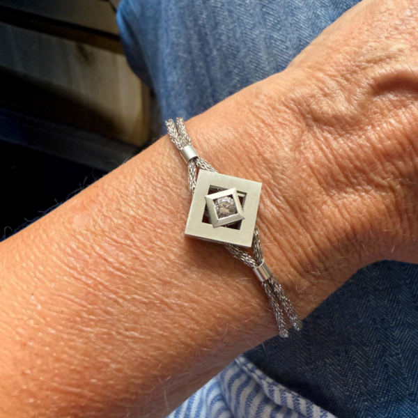 bracelet mikado argent zircon porte | 26 Passage, Créatrice de bijoux à Paris Bracelet with a braided silver chain and a square concentric pendant featuring a central diamond on a wrist.