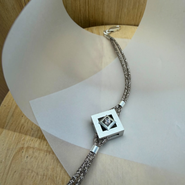bracelet mikado argent zircon vue de haut | 26 Passage, Créatrice de bijoux à Paris Silver multi-strand necklace with a square pendant featuring a central gemstone on a curved white display board.