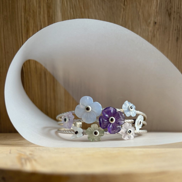 bracelet prairie pierres en argent | 26 Passage, Créatrice de bijoux à Paris Pastel flower bracelet with blue, purple, pink, and green blossom charms on a braided cord, resting on a curved white surface.
