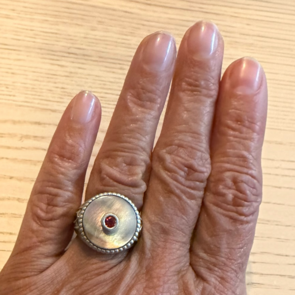chevaliere nacre saphir sonkia portee | 26 Passage, Créatrice de bijoux à Paris Close-up of a hand wearing a silver circular ring with a small red gemstone on the middle finger, textured band visible on a wooden surface