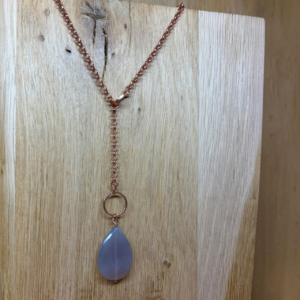 collier lasso vermeil et agate grise forme poire Rose‑gold chain necklace with a blue teardrop pendant hanging from a circular ring, set against a wooden background.