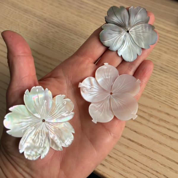 Hand holding four carved flower-shaped resin buttons in pearly white and iridescent finishes on the palm of a hand.
